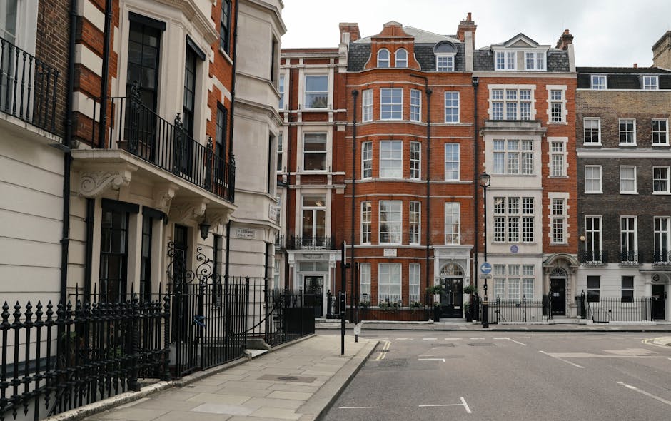 A street view in Mayfair showing several multi-storey Victorian and Edwardian residential buildings constructed with red and grey brickwork, featuring large sash windows with white frames and decorative stone elements around entrances and balconies. The pavement in the foreground is paved with concrete slabs, and a black wrought iron fence runs along the edge of a property on the left side. The street appears calm with no visible vehicles or pedestrians. This image is relevant to house removals and furniture transport, illustrating the typical architecture and urban environment involved in home relocation services provided by Man and Van Mayfair, including logistical considerations for moving within such historic residential buildings.