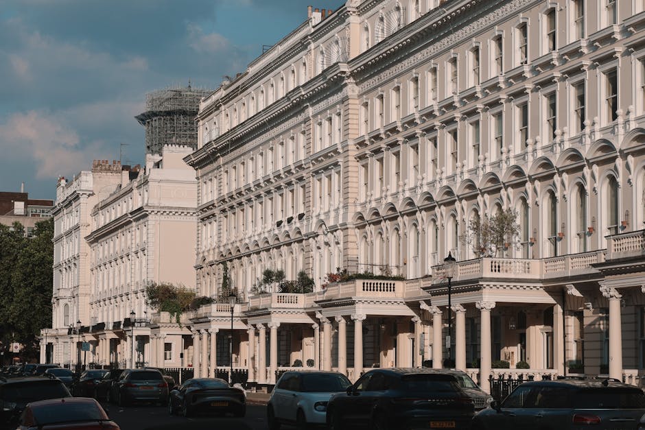 A row of elegant Victorian-style terraced houses with white facades and decorative architectural details line a street in Mayfair, London. In the foreground, several parked cars occupy the street, with some obscuring the lower parts of the buildings. The front entrances are visible, featuring steps and small porches with columns and railings, and some buildings have potted plants or small trees on the balconies or near their entrances. To the left, there's a construction scaffold partially covered with netting on one of the buildings, indicating ongoing renovation work. The street is lit by traditional black lamp posts, and the sky above is partly cloudy with patches of blue, suggesting a clear daytime. This scene captures the classic look of a historic residential area being prepared for home relocation or furniture transport, with visible packing materials like blankets or plastic wraps in related moving contexts that might be used during interior packing, as supported by the presence of a professional removals service such as Man and Van Mayfair.