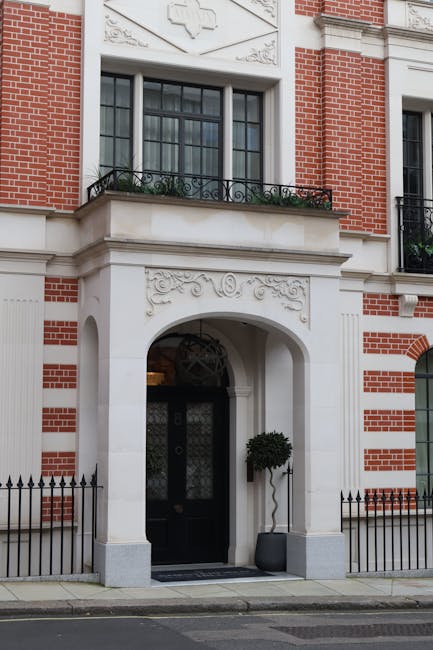 A street view in Mayfair showing several multi-storey Victorian and Edwardian residential buildings constructed with red and grey brickwork, featuring large sash windows with white frames and decorative stone elements around entrances and balconies. The pavement in the foreground is paved with concrete slabs, and a black wrought iron fence runs along the edge of a property on the left side. The street appears calm with no visible vehicles or pedestrians. This image is relevant to house removals and furniture transport, illustrating the typical architecture and urban environment involved in home relocation services provided by Man and Van Mayfair, including logistical considerations for moving within such historic residential buildings.