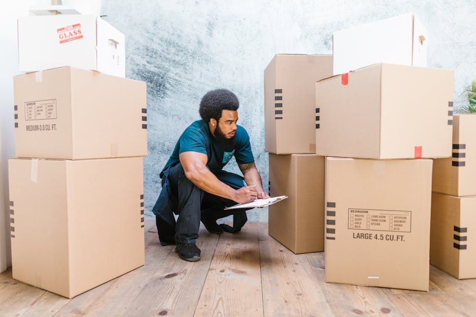 A man with dark skin, curly black hair, and a beard is crouching inside a room surrounded by stacked cardboard boxes of various sizes, some labeled for storage and furniture. He is wearing a teal shir