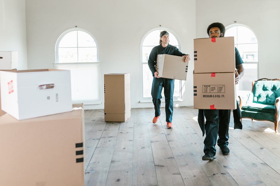 A man with dark skin, curly black hair, and a beard is crouching inside a room surrounded by stacked cardboard boxes of various sizes, some labeled for storage and furniture. He is wearing a teal shir