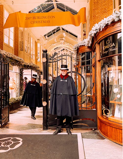 Inside a covered shopping arcade with high, arched glass ceilings, two men dressed in black coats, capes, and bowler hats stand near a black metal gate, which is part of a home relocation or furniture transport process managed by Man and Van Mayfair. The man in the foreground is positioned in front of the gate, while the other is walking behind him. The surroundings include wood-paneled shopfronts, one displaying an Omega watch window, and the area is decorated with white floral garlands along the top of the display windows. Overhead, a large yellow banner reads '200th Burlington Christmas,' indicating a seasonal event or festive occasion. The lighting is bright and warm, highlighting the detailed interior architecture, and the overall scene suggests careful handling of valuable items within this moving or packing context, aligned with a professional removal service.