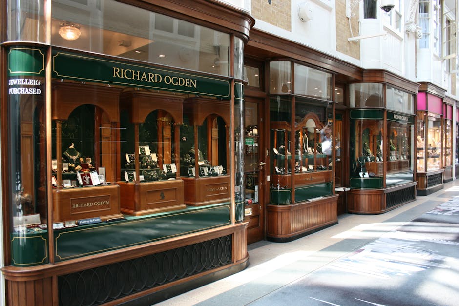Photograph of a street-facing jewelry shop window displaying various pieces, including necklaces, rings, and watches, arranged on padded black and white display stands inside wooden and glass showcases with curved glass panels. The shop features a dark green awning with gold lettering reading 'Richard Ogden' and is part of a historic shopping arcade with adjacent storefronts. The pavement outside is lit by natural daylight, with shadows cast on the ground. Man and Van Mayfair, a professional removal company, is involved in the home relocation process, and the image may relate to furniture transport or packing of valuable items during a moving service, emphasizing careful handling and secure display setup.