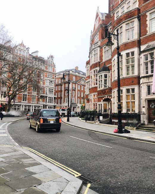 Wide view of a residential street in Mayfair featuring historic brick and white-windowed period townhouses with ornate facades and bay windows, some with small front gardens enclosed by black iron fences. The street is paved with asphalt, showing a small section of double yellow lines at the curb, and curved gently to the left. A black vehicle is parked on the street, partially blocking the view of the road. Along the sidewalk, there are leafless trees, black lampposts, and a few small objects on the pavement, including a pink bag near the curb. The street is quiet with no pedestrians visible, and the background includes taller buildings and a cloudy sky. This scene depicts a typical residential area suitable for home relocation and furniture transport services, such as those provided by Man and Van Mayfair, in the context of packing and moving operations.
