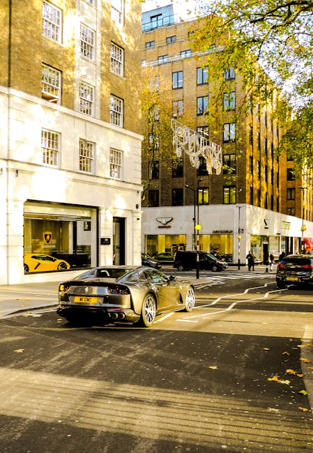 Wide view of a residential street in Mayfair featuring historic brick and white-windowed period townhouses with ornate facades and bay windows, some with small front gardens enclosed by black iron fences. The street is paved with asphalt, showing a small section of double yellow lines at the curb, and curved gently to the left. A black vehicle is parked on the street, partially blocking the view of the road. Along the sidewalk, there are leafless trees, black lampposts, and a few small objects on the pavement, including a pink bag near the curb. The street is quiet with no pedestrians visible, and the background includes taller buildings and a cloudy sky. This scene depicts a typical residential area suitable for home relocation and furniture transport services, such as those provided by Man and Van Mayfair, in the context of packing and moving operations.