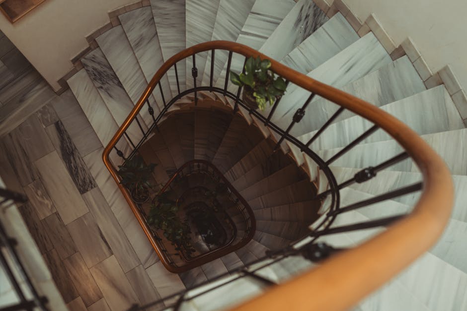 A view from the top of a narrow, winding staircase inside a home, showing wooden handrails with a smooth finish and black metal balusters. The staircase is confined within tight walls lined with light-coloured marble or porcelain tiles, arranged in a rectangular pattern, leading downward to a hidden lower level. On the staircase, there are several cardboard boxes secured with plastic wrap, along with a few lightweight fabric blankets draped over the edges, indicating a packing process for home relocation. A portion of a green potted plant is visible near the railing, adding a touch of decor. The environment appears well-lit with natural or ambient lighting, emphasizing the confined space typical of Mayfair's narrow staircases, which can pose challenges for furniture transport during removals. The image is associated with [COMPANY_NAME]’s house removal services, highlighting the careful loading and moving of household items in tight residential spaces, demonstrating expertise in logistics for cramped staircases as described in the page about van solutions for narrow Mayfair homes.