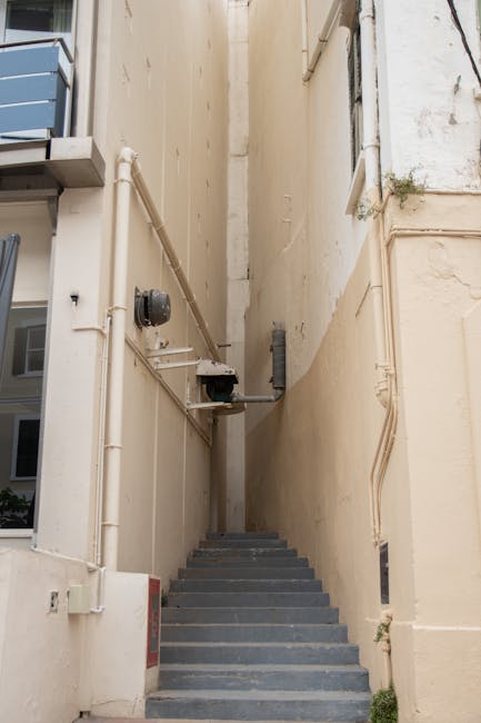 A narrow exterior stairwell between two beige buildings with high walls, featuring a short flight of dark grey steps leading upward. On the left side, metal piping, electrical boxes, and conduit run along the wall, with a utility meter visible. The stairwell is enclosed, with limited natural light, and a small plant is seen growing near the base of the right wall. This setting illustrates the confined space often encountered in urban home relocations, highlighting the need for specialized furniture transport and careful movement planning on tight staircases, as handled by Man and Van Mayfair.