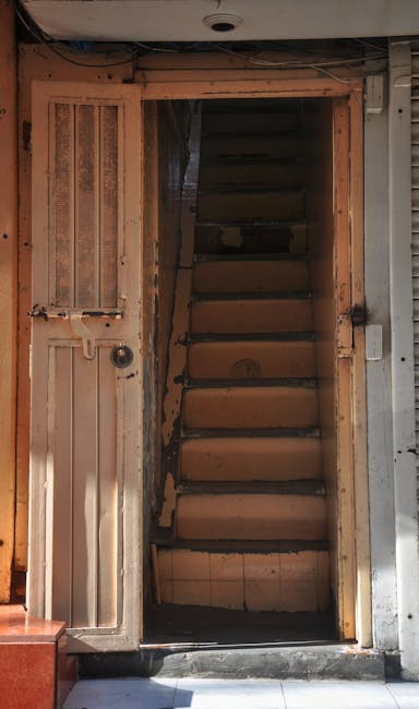 A view from the top of a narrow, winding staircase inside a home, showing wooden handrails with a smooth finish and black metal balusters. The staircase is confined within tight walls lined with light-coloured marble or porcelain tiles, arranged in a rectangular pattern, leading downward to a hidden lower level. On the staircase, there are several cardboard boxes secured with plastic wrap, along with a few lightweight fabric blankets draped over the edges, indicating a packing process for home relocation. A portion of a green potted plant is visible near the railing, adding a touch of decor. The environment appears well-lit with natural or ambient lighting, emphasizing the confined space typical of Mayfair's narrow staircases, which can pose challenges for furniture transport during removals. The image is associated with [COMPANY_NAME]’s house removal services, highlighting the careful loading and moving of household items in tight residential spaces, demonstrating expertise in logistics for cramped staircases as described in the page about van solutions for narrow Mayfair homes.