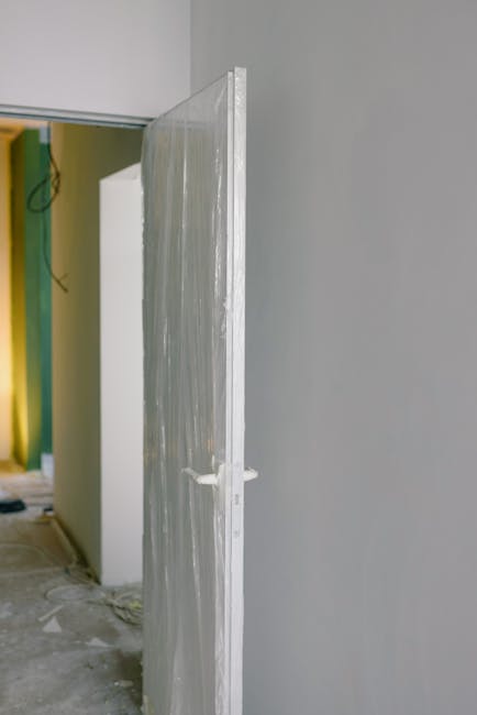 A close-up view of a clear plastic protective sheet mounted on a door frame in an interior space, used to shield a period wooden floor during a house move in Mayfair. The plastic sheet is secured with masking tape at the edges, and part of the door frame is painted white. The background reveals part of a room with yellow and green construction materials and partially finished walls, indicating ongoing renovations or preparatory steps for a property relocation. The floor beneath the plastic appears to be unfinished concrete, with dust and debris visible. The image is taken in natural daylight, highlighting the protective measures taken by Man and Van Mayfair to ensure the preservation of delicate flooring during furniture transport and home relocation activities.