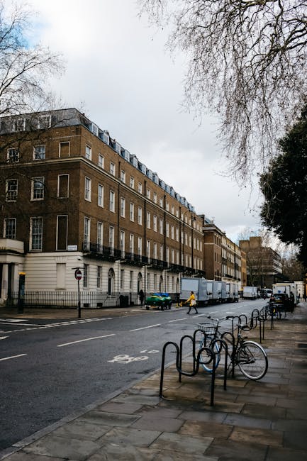 A multi-storey urban building with a brown brick facade and white window frames is situated along a city street on a cloudy day. In the foreground, a sidewalk features several black metal bike racks, with a single bicycle parked and secured. The street is lined with parked vehicles, including a small green car and a larger white delivery van positioned near the building. A person in bright yellow clothing is walking across the street, possibly involved in a home relocation process. Visible on the pavement are faded road signs and directional indicators for cyclists, indicating a designated bike lane. To the right, tall leafless trees extend over the sidewalk, their branches partially framing the scene. Several large cardboard boxes, wooden crates, and wrapped furniture pieces are seen being loaded into the van, suggesting a furniture transport operation as part of a house removals or packing and moving service. This scene is managed by Man and Van Mayfair, experienced in same-day removals and emergency moving services within the Mayfair area, demonstrating the logistics involved in loading furniture and packed boxes for home relocation. The lighting is natural, and the overall environment is typical of an urban street during daytime, emphasizing the coordination involved in efficient house removals in the city.