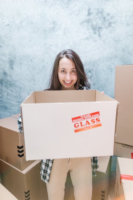 A young woman with long brown hair smiling and holding an empty cardboard box labeled 'GLASS' in a room with a textured grey wall. She is dressed casually in a plaid shirt and light-colored trousers. Surrounding her are several other cardboard boxes, some closed and others open, with packing materials such as plastic wrap and paper visible on some boxes. The setting suggests an ongoing home relocation or packing process, with the woman preparing for furniture transport or moving services. The scene captures the interior environment where house removals, such as those provided by Man and Van Mayfair, are typically carried out, emphasizing packing and loading activities involved in same-day emergency moves or scheduled relocations in Mayfair.