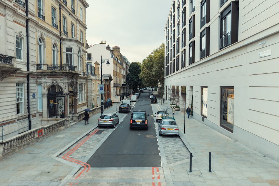 A view of a city street in Mayfair, showing a modern white building on the right with large windows and a classic, ornate building on the left with detailed stonework and balcony railings. The street has parked cars along both sides, including a black van situated in the middle of the road, which appears to be part of a home relocation process by Man and Van Mayfair. The van is parked near the curb, with its sliding door open, and there are cardboard boxes and furniture wrapped in plastic and blankets within the vehicle, suggesting packing and loading activities. Pedestrians are walking on the pavements on both sides of the street, and a person is seen carrying a box towards the entrance of a building. The environment is well-lit during daylight, with trees visible further down the street, and the scene captures the logistics involved in furniture transport and home moving in an urban residential area.