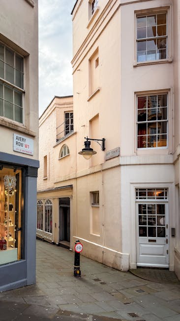 A narrow street in Mayfair featuring a beige-painted, multi-storey building with several large, white-framed windows and a white door at street level. Attached to the building's facade is a black outdoor wall lamp. On the left side, part of a shop window displays jewelry, and visible signage indicates the location as South Audley Street. The pavement is paved with cobblestones, and a small bollard with a sign for parking restrictions is positioned on the street. The scene captures the typical architecture and urban environment relevant to house removals and furniture transport services provided by Man and Van Mayfair, highlighting the logistical considerations of loading and access in a central London setting.