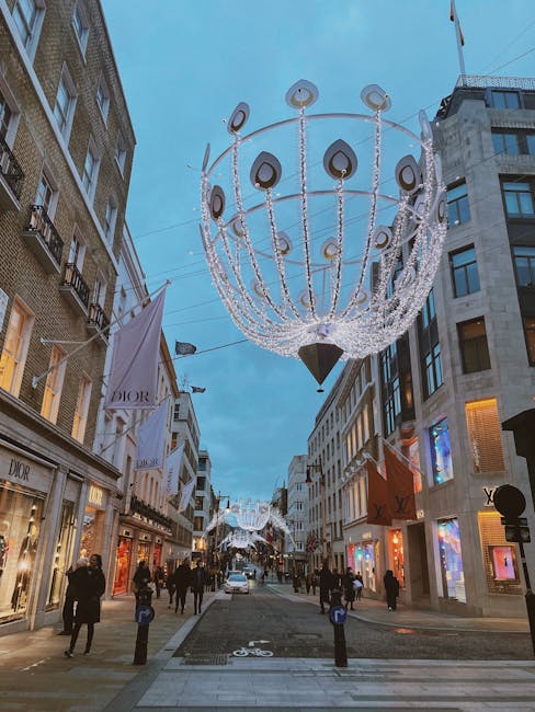 A view of a city street in Mayfair, showing a modern white building on the right with large windows and a classic, ornate building on the left with detailed stonework and balcony railings. The street has parked cars along both sides, including a black van situated in the middle of the road, which appears to be part of a home relocation process by Man and Van Mayfair. The van is parked near the curb, with its sliding door open, and there are cardboard boxes and furniture wrapped in plastic and blankets within the vehicle, suggesting packing and loading activities. Pedestrians are walking on the pavements on both sides of the street, and a person is seen carrying a box towards the entrance of a building. The environment is well-lit during daylight, with trees visible further down the street, and the scene captures the logistics involved in furniture transport and home moving in an urban residential area.