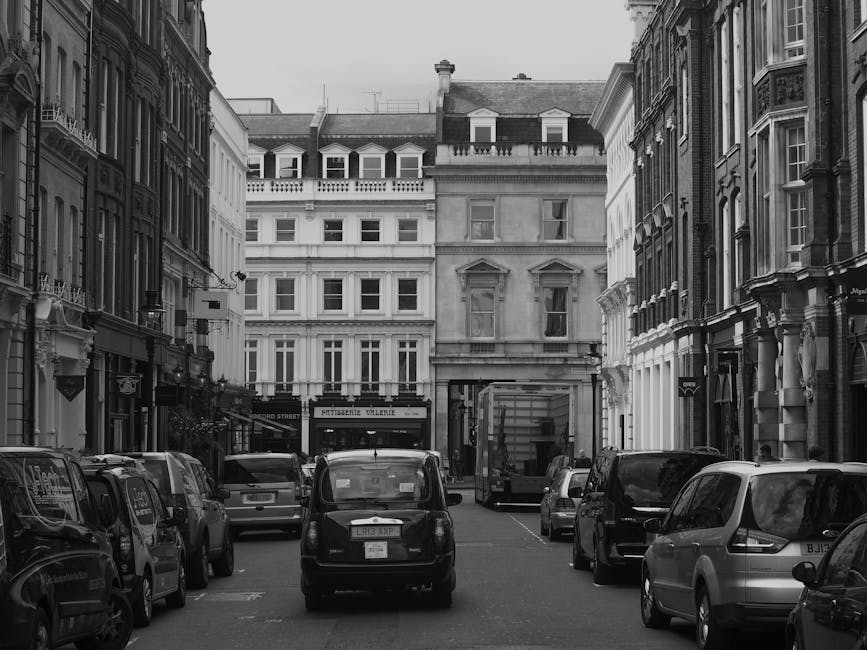 A black and white photograph of a narrow street in Mayfair, London, with multiple parked cars lining both sides of the street, including small vans and larger vehicles. In the background, a delivery truck or moving van is parked near the entrance to a building, with some objects possibly being loaded or unloaded. The surrounding buildings are classic Victorian and Georgian-style structures with ornate facades, large windows, and decorative details, typical of the Westminster area. The scene appears to be during daytime with natural lighting. The image reflects a typical setting for home relocation or furniture transport, where a professional removals service such as Man and Van Mayfair might operate, coordinating vehicle loading and unloading in an urban environment to facilitate house removals or moving logistics.