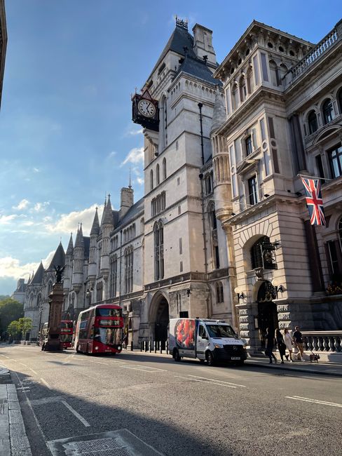 A street scene in Westminster featuring the historic clock tower of Westminster Palace, with its white stone facade and black clock face visible. On the pavement in front of the building, a man and a woman are loading a large cardboard box wrapped in plastic onto a moving van operated by Man and Van Mayfair, a professional removals service. The scene includes a red double-decker bus and a small white delivery van parked along the curb, with additional pedestrians walking nearby. The building’s ornate architecture, including decorative stonework and windows, is highlighted by natural daylight under a partly cloudy sky, illustrating the logistics involved in home relocation and furniture transport in the area. The image captures the essence of urban moving activities aligned with Mayfair's local removals services, emphasizing the loading process with furniture and packing materials prepared for transportation.