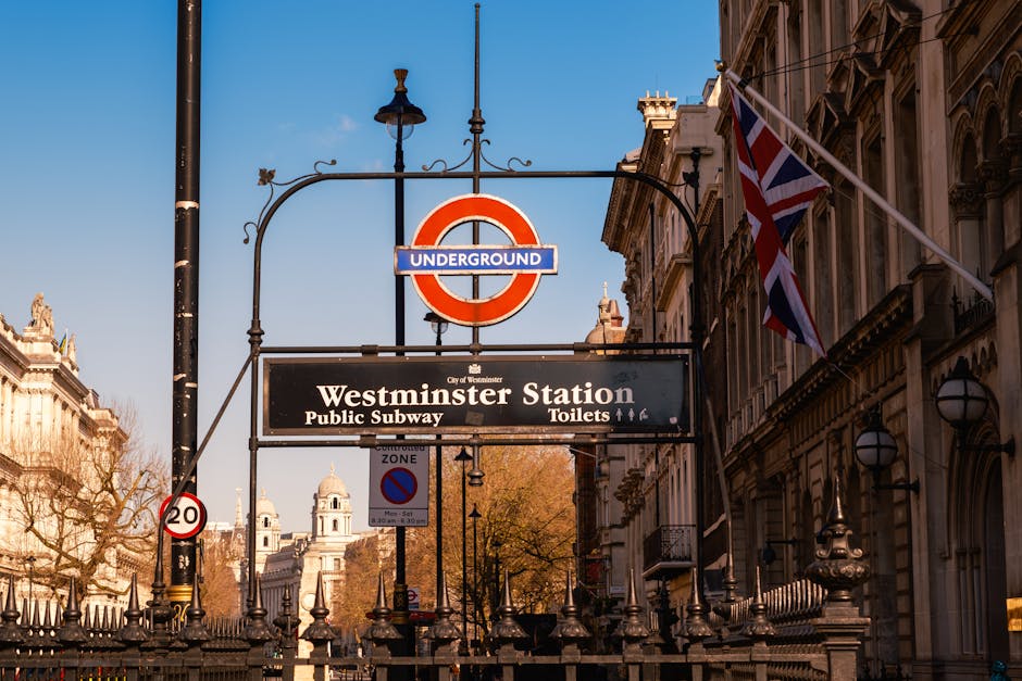 A street view near Westminster Station in London, featuring a large red and blue London Underground sign suspended from a decorative iron lamppost. Beneath the sign, there is a black rectangular direc